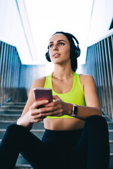 Contemplative female runner in electronic technology resting at urban stairs and thinking about ABT...