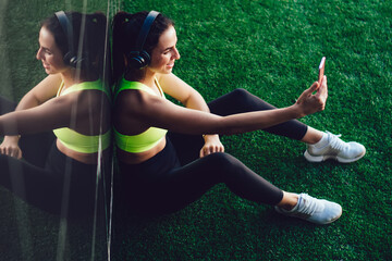 Happy youth sportswoman using cellular device for shooting influence video vlog during workout break, millennial athlete in electronic headphones calling to friend via application for communicate