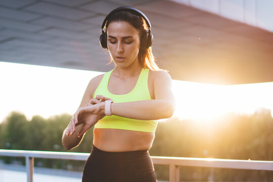Young Caucasian female jogger in headphones enjoying music playlist using wearable device on wrist for GPS and time tracking on workout training, sporty woman in earphones looking at smartwatch