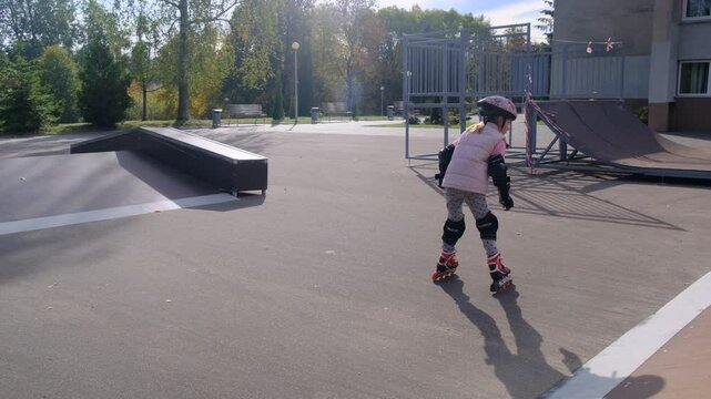 A young girl, wearing a helmet and protective gear, prepares to roller skate. She is poised and ready for action on a smooth, paved path, with autumnal trees in the background
