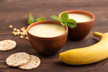 Organic non dairy banana and soy milk in clay cup on brown wooden, side view, selective focus.