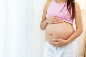 Close up pregnant woman stand with touch her belly in front of white curtain look relax with happiness.