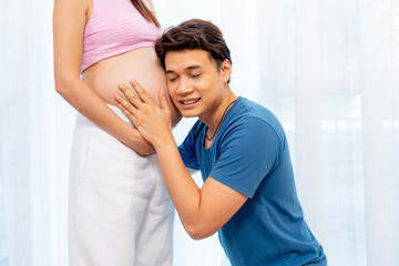 Naklejka premium Asian man sit down and attach ear to belly of pregnant woman with smiling to express love and care in front of white curtain in their house.