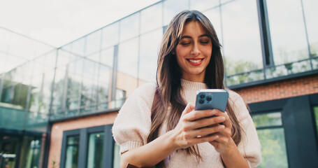 Cute Caucasian woman using her mobile device. Standing in front of college or university. Typing on...
