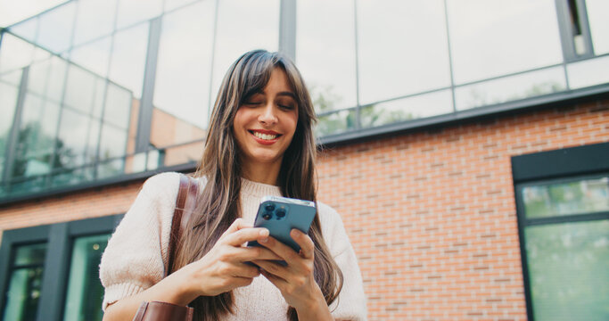 Camera front view of happy Caucasian girl walking away from modern college. Student walking back home. Using her smartphone with both hands. Texting. Typing message on touch screen.