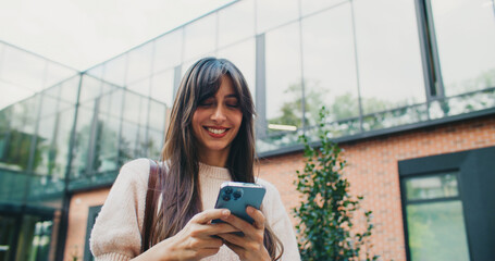 Front camera view of young teachers assistant walking home after working day. Smiling while chatting with her husband on smartphone. Reading positive message on screen of mobile device.