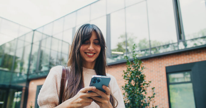 Front camera view of young teachers assistant walking home after working day. Smiling while chatting with her husband on smartphone. Reading positive message on screen of mobile device.