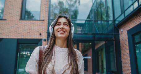 Charming Caucasian female looking straight ahead while smiling. Placing headphones on head and starting listening to video or stream online. Walking away from office building with big windows.