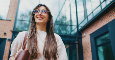 Positive Caucasian woman confidently walking away from office building. Leaving work early. Smiling while looking around. Happy about upcoming weekend. Wearing round glasses and handbag.