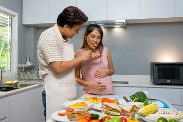 Asian man give a piece of fruit to pregnant woman and look each other with love and care stay in the kitchen of their house.