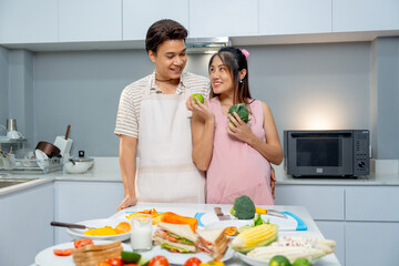 Asian man stand beside pregnant woman who hold apple and vegetable and express love and care stay in the kitchen of their house.