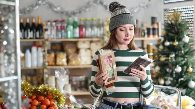 While shopping on eve of New Year holidays, young lady buys sweets for festive party. Buyer examines packages with sweet dessert snacks, purchases chocolates and panellets, nougat. 