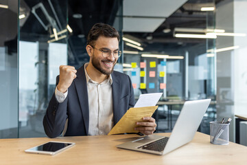 A happy businessman celebrates after reading good news in an office. He holds a letter and displays...