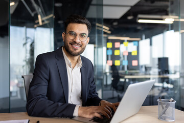 Fototapeta premium A confident businessman in a suit is working on a laptop in a bright, modern office. The office features glass walls and colorful sticky notes, creating an open and collaborative work environment.