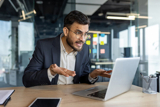 An exasperated businessman in an office uses a laptop for a video call, expressing frustration. His smartphone is visible, adding to the tech-focused environment.