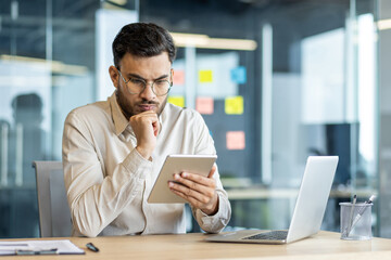 Obraz premium A business professional intensely studies information on a digital tablet in a contemporary office. The setting is characterized by a modern, tech-savvy atmosphere