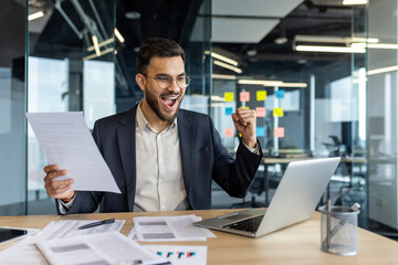 A cheerful businessman in a suit celebrates a successful achievement at his office desk. The image captures a moment of joy and motivation while reviewing documents on a laptop.