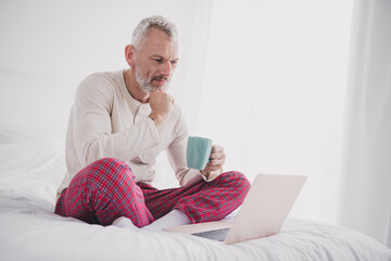 Mature man enjoying a relaxed morning on bed with coffee and laptop in cozy pajamas