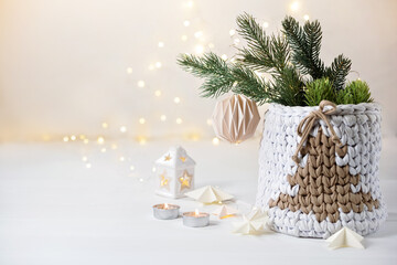 Cozy Christmas decoration. Knitted basket with Christmas tree, paper ornament and lights on festive lights background. Hygge atmosphere