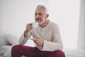 Cheerful mature man enjoys a morning coffee in pajamas, embracing tranquility in a cozy bedroom setting