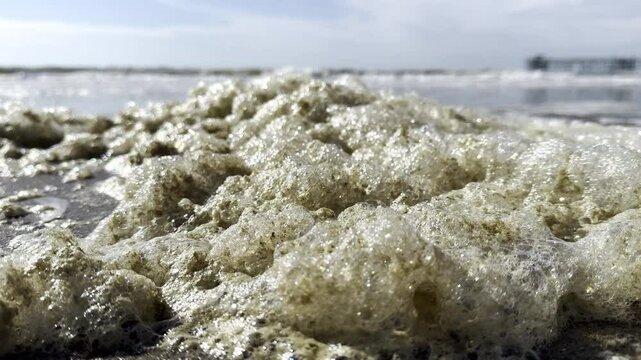 Piles of sea foam jiggling in the wind with waves in the distance seen from a low angle on Oak Island, North Carolina