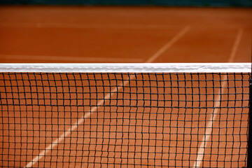 Close-up of a tennis net over an orange clay court.  The net's black mesh and white top are clearly...