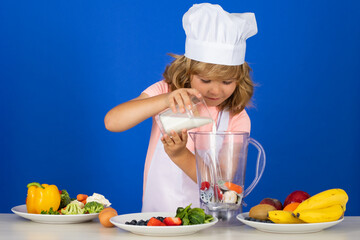 Portrait of a 7, 8 years old child in cook cap and apron making milk shake smoothie and cooking food in kitchen. Cute little blonde happy smiling chef.