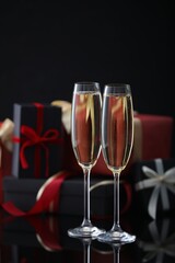Delicious champagne in glasses and Christmas presents on black mirror surface