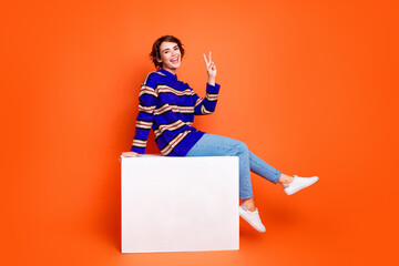 Full body length photo of brown hair woman in trendy outfit sitting white podium peaceful v sign isolated on orange color background