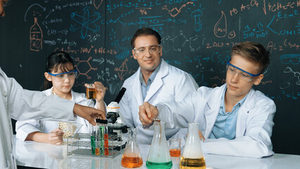 Creative teacher pointing chemistry at blackboard while talking to highschool boy at table with microscope and test tube with colored liquid. Young student wearing lab coat in STEM class. Edification.