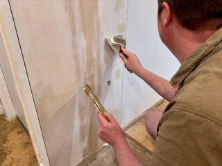 A man uses a putty knife and plaster to smooth a wall seam on plasterboard, preparing the surface for painting during a DIY home renovation project.
