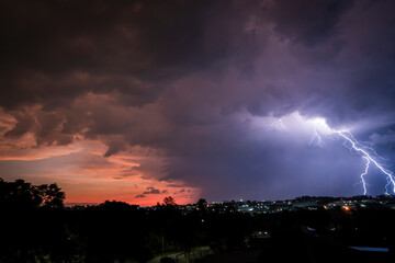 A stunning photograph of a lightning strike over a suburban area during a vibrant sunset, featuring dramatic orange and pink hues against a moody sky. Striking in between houses and factories