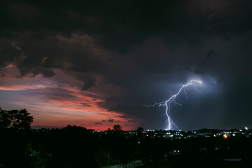 A stunning photograph of a lightning strike over a suburban area during a vibrant sunset, featuring dramatic orange and pink hues against a moody sky. Striking in between houses and factories