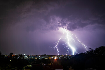 A stunning photograph of a lightning strike over a suburban area during a vibrant sunset, featuring dramatic orange and pink hues against a moody sky. Striking in between houses and factories © Phillip