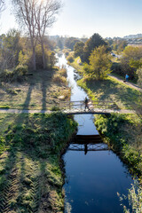 Fototapeta premium Sunlight casts long shadows over a serene stream landscape from the Haagse Beek, featuring a small bridge and lush greenery. A person stands on the bridge, reflecting in the calm water below.