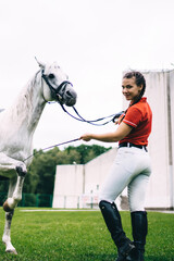 Obraz premium Full length portrait of Caucasian jockey girl posing during dressage competition at nature, female horseback rider looking at camera while white champion stallion animal showing stunt outdoors