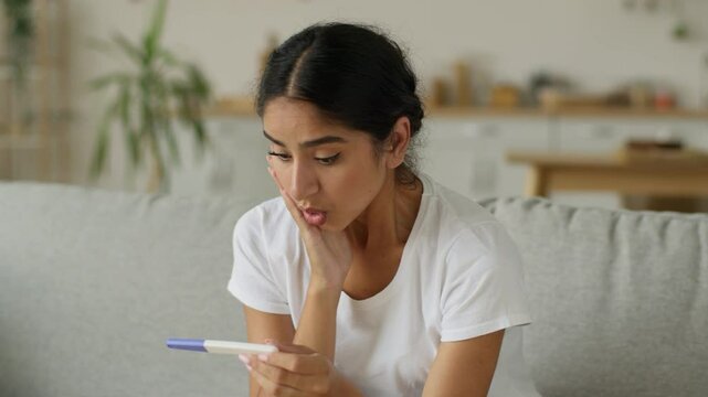 Shocked puzzled confused amazed wonder Indian woman Arabian lady female girl holding positive result pregnancy test shock unwanted unplanned pregnant at home couch medicine health childbirth wait baby