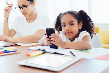 Mother using laptop and daughter with phone at table in living room