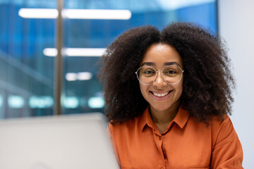 A confident young professional woman wearing glasses and an orange shirt, smiling while sitting at a desk in a modern office setting. She appears composed and approachable.