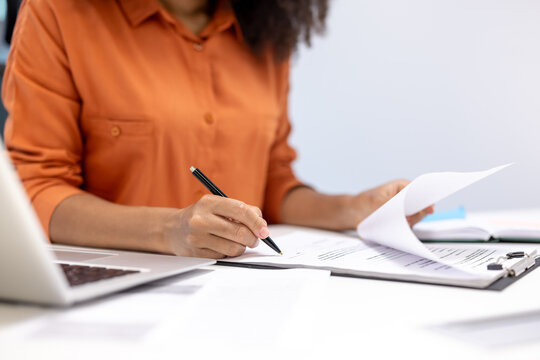 A professional woman in an orange blouse is signing important documents at her desk. The image captures a focus on her hands and the paperwork, suggesting productivity and business activities.