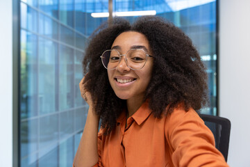 A cheerful young woman with glasses takes a selfie at her office desk. Natural lighting and contemporary design elements enhance the friendly atmosphere depicted in this professional setting.