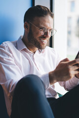 Happy male blogger in optical spectacles for provide eyes protection enjoying smartphone communication with virtual followers from social networks, cheerful man typing content text on cellphone