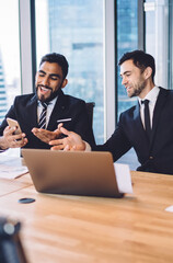 Happy multicultural male colleagues smiling during brainstorming meeting using smartphone device for browsing website for networking, cheerful business consultants enjoying friendship at job