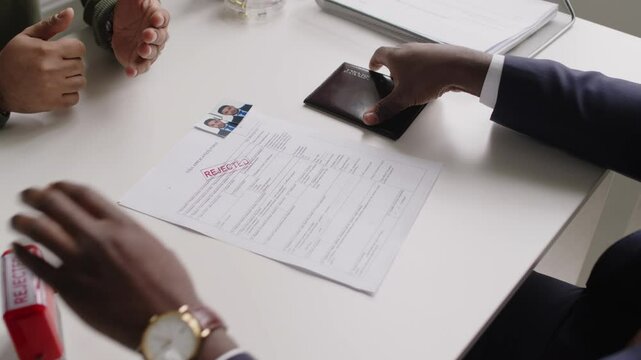 High angle shot of hands of Black male consular officer putting Rejected stamp on applicants visa documents during interview in embassy