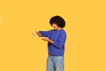 A young black man stands against a vibrant yellow backdrop, joyfully interacting with cash in his hands. His casual outfit reflects a modern lifestyle, showcasing a moment of enthusiasm.