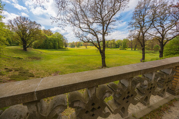 Beautiful landscape of big green park with green glades, trees at cloudy sunny morning seen from small bridge with concrete fence of in park