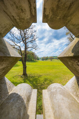 Beautiful landscape of big green park with green glades, trees at cloudy sunny morning seen through decorative hole in concrete fence of small bridge in park