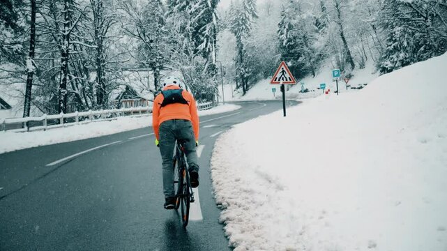 A male cyclist rides up a winding road in the mountains as snow falls