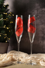Christmas decor, two transparent glass with a red ribbon inside, tree with lights, dark background, beige cloth on wooden table