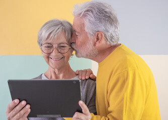 Caucasian smiling modern senior woman looking at digital tablet, husband kissing her expressing love and affection